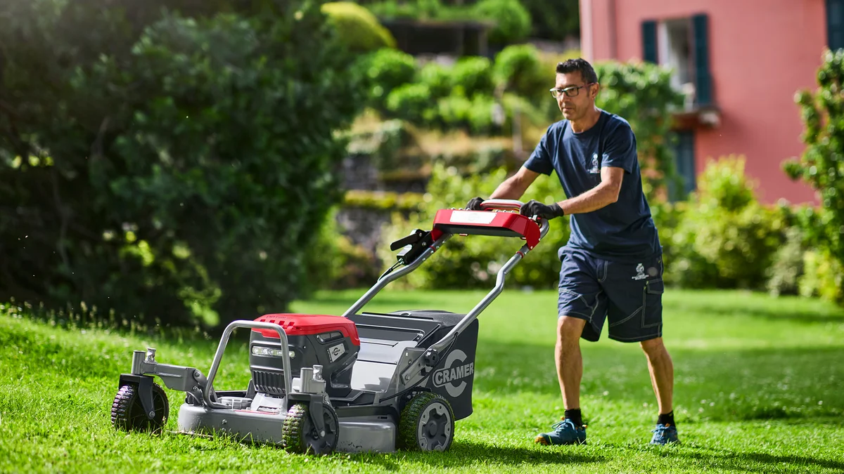 Homme poussant la tondeuse à conducteur marchant Cramer ATM76 rouge et grise sur une pelouse verte, jardin avec maison en arrière-plan